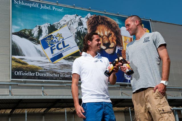 Die beiden FCL Spieler Adrian Winter (lin ks) und Daniel Fanger posieren mit dem FCL-Fan-Bier vor dem riesigen FCL Plakat an der Fassade der Brauerei Eichhof. (Bild: Roger Grütter/Neue LZ)