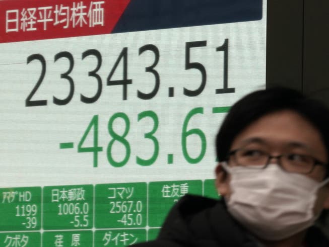 An den Börsen geht es wegen dem Coronavirus abwärts: ein Mann mit einer Maske in Tokio vor einer Börsenanzeigetafel.