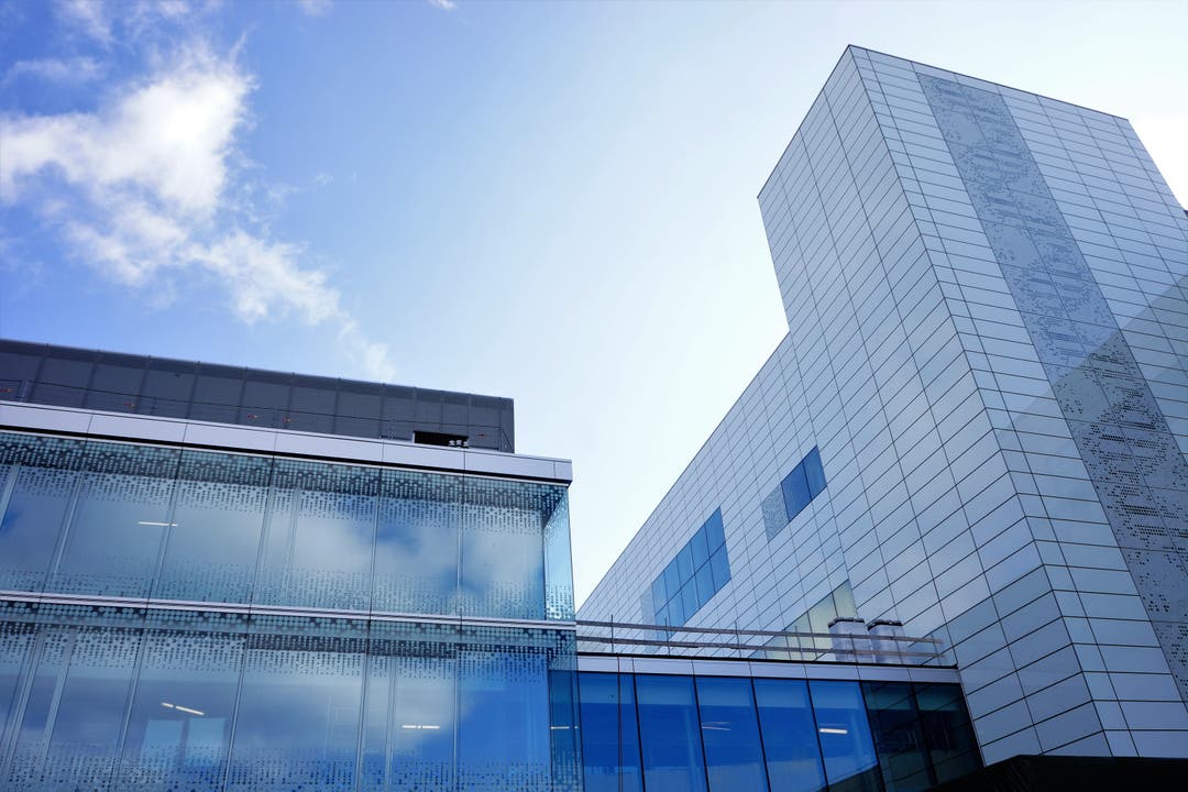 Die Hülle des Hauptgebäudes spiegelt das aktuelle Wetter. Links ist ein Teil des Empfangs- und Administrativgebäudes. Rechts ist ein Teil des Hauptgebäudes mit der Doppelhelix, die an die DNA-Struktur erinnert, in der Glasfassade des Treppenhauses.