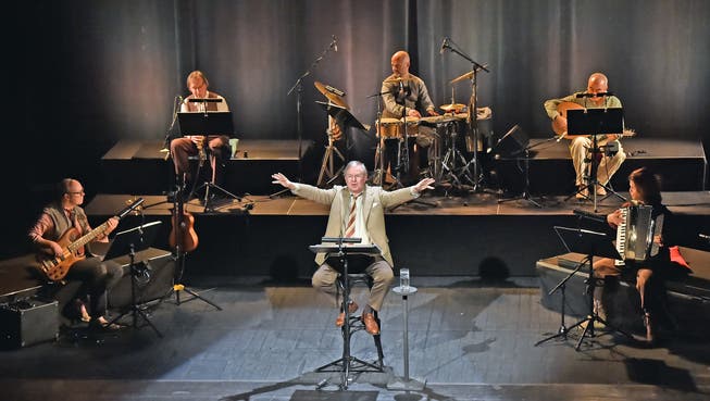 Joachim Król auf einem Barhocker; hinter und neben sich das fünfköpfige L’Orchestre du Soleil.
