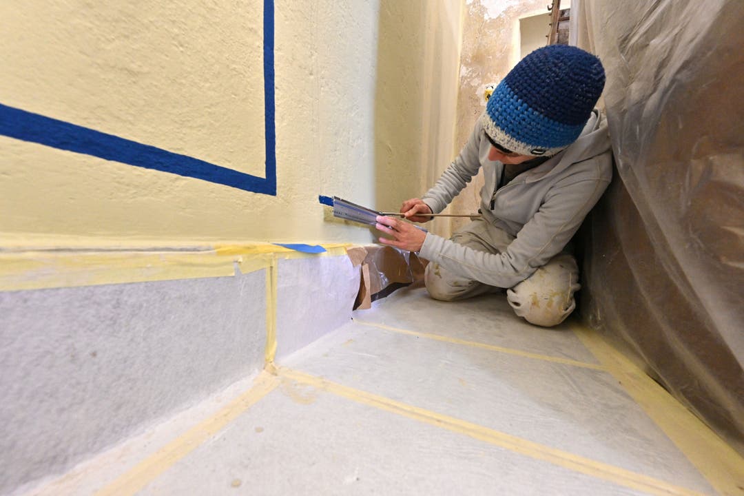 Blaue Zierleisten in der Bauernstube. Eine Malerin streicht die Farbe entlang des Lineals auf die Wandoberfläche.
