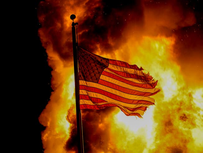 Eine ausgefranste US-Flagge weht vor den Flammen eines Feuers bei einem Protest nach den Schüssen auf Jacob Blake. Foto: Morry Gash/AP/dpa