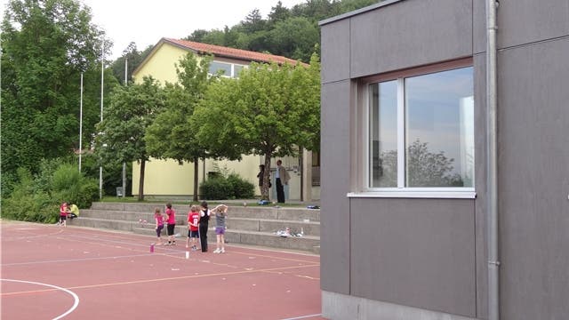 Das bestehende Schulhaus (links, Teilansicht) soll durch einen Anbau ergänzt werden; rechts die Teilansicht des aufgestellten provisorischen Schulraums. – Foto: chr