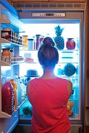 A young caucasian woman standing in front of the open refrigerator at late night, contemplating and wondering about a midnight snack in a domestic home kitchen. She is dressed in a bath robe hungry and looking for food. A symbol of dieting lifestyle. Photographed in vertical format. (Bild: YinYang (E+))