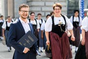 Elias Räber posiert am Bahnhof Luzern mit seiner Masterarbeit, während eine Trachtengruppe vorbeiläuft. (Bild: Eveline Beerkircher, 14. September 2019)