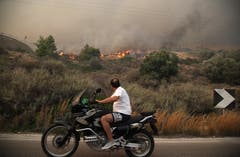 Ein Motorradfahrer schaut in Kineta, westlich von Athen, auf brennende Büsche. (Bild: AP Photo/Thanassis Stavrakis (23. Juli 2018))